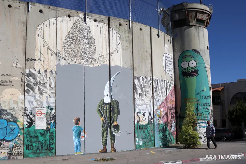 A Palestinian man walks past a graffiti mural depicting a boy holding a fishbowl looking at a uniformed Israeli soldier wearing a white hood in the style of the Ku Klux Klan (KKK), drawn by Palestinian artist Taqi Spatee...