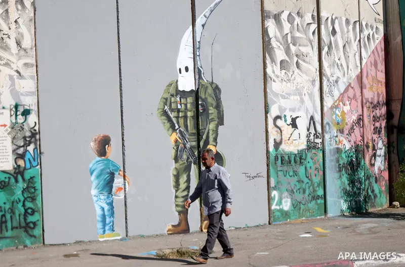 A Palestinian man walks past a graffiti mural depicting a boy holding a fishbowl looking at a uniformed Israeli soldier wearing a white hood in the style of the Ku Klux Klan (KKK), drawn by Palestinian artist Taqi Spatee...