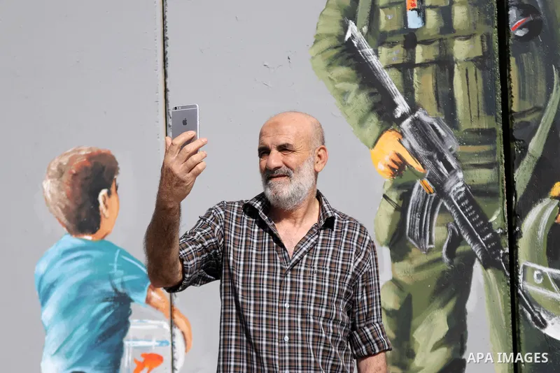 A man uses his phone to take a "selfie" photo near a graffiti mural depicting a boy holding a fishbowl looking at a uniformed Israeli soldier wearing a white hood in the style of the Ku Klux Klan (KKK), drawn by Palestin...