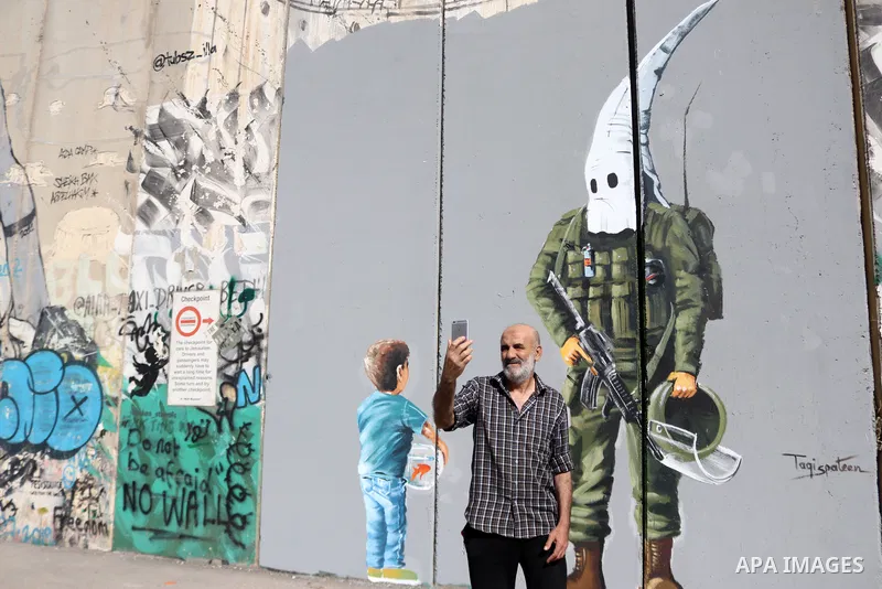A man uses his phone to take a "selfie" photo near a graffiti mural depicting a boy holding a fishbowl looking at a uniformed Israeli soldier wearing a white hood in the style of the Ku Klux Klan (KKK), drawn by Palestin...