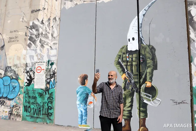 A man uses his phone to take a "selfie" photo near a graffiti mural depicting a boy holding a fishbowl looking at a uniformed Israeli soldier wearing a white hood in the style of the Ku Klux Klan (KKK), drawn by Palestin...