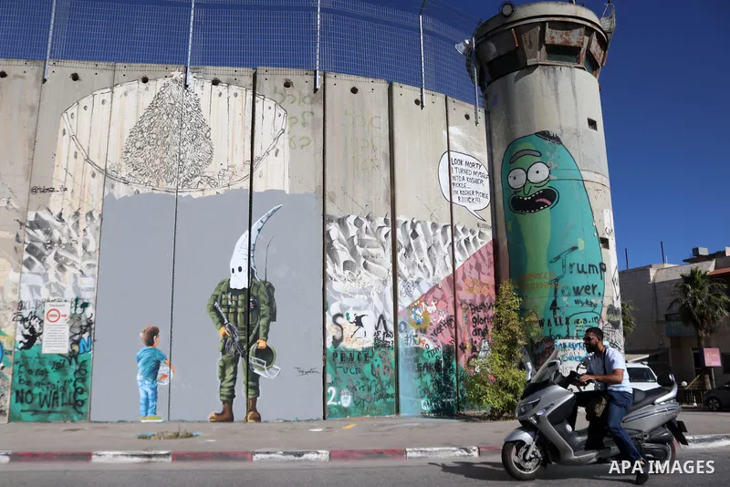 Palestinians walk past a graffiti mural depicting a boy holding a fishbowl looking at a uniformed Israeli soldier wearing a white hood in the style of the Ku Klux Klan (KKK), drawn by Palestinian artist Taqi Spateen alon...