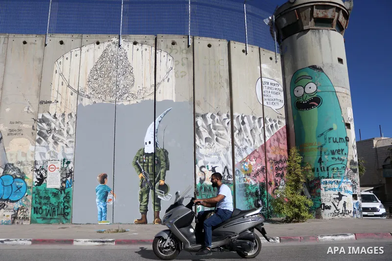 Palestinians walk past a graffiti mural depicting a boy holding a fishbowl looking at a uniformed Israeli soldier wearing a white hood in the style of the Ku Klux Klan (KKK), drawn by Palestinian artist Taqi Spateen alon...