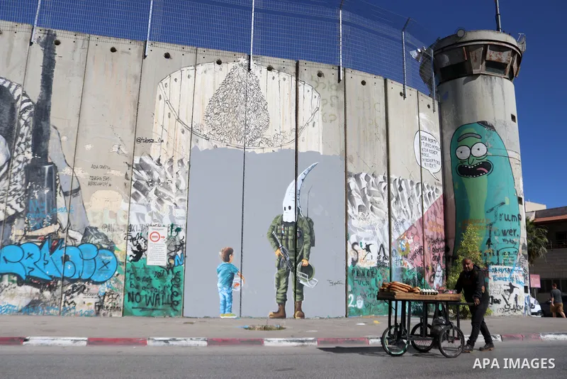 A street food vendor pushes his cart past a graffiti mural depicting a boy holding a fishbowl looking at a uniformed Israeli soldier wearing a white hood in the style of the Ku Klux Klan (KKK), drawn by Palestinian artis...