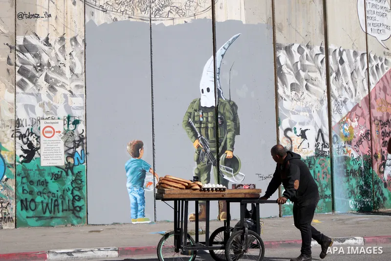 A street food vendor pushes his cart past a graffiti mural depicting a boy holding a fishbowl looking at a uniformed Israeli soldier wearing a white hood in the style of the Ku Klux Klan (KKK), drawn by Palestinian artis...