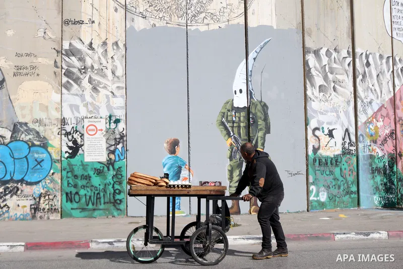 A street food vendor pushes his cart past a graffiti mural depicting a boy holding a fishbowl looking at a uniformed Israeli soldier wearing a white hood in the style of the Ku Klux Klan (KKK), drawn by Palestinian artis...