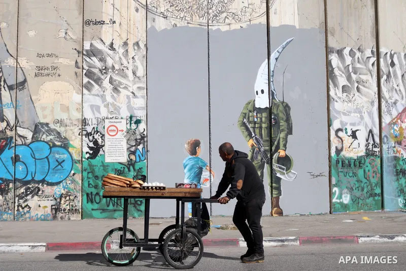 A street food vendor pushes his cart past a graffiti mural depicting a boy holding a fishbowl looking at a uniformed Israeli soldier wearing a white hood in the style of the Ku Klux Klan (KKK), drawn by Palestinian artis...