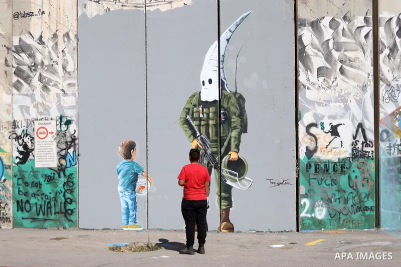 A Palestinian man walks past a graffiti mural depicting a boy holding a fishbowl looking at a uniformed Israeli soldier wearing a white hood in the style of the Ku Klux Klan (KKK), drawn by Palestinian artist Taqi Spatee...