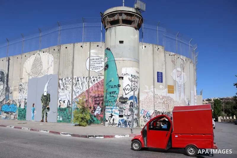 Palestinians walk past a graffiti mural depicting a boy holding a fishbowl looking at a uniformed Israeli soldier wearing a white hood in the style of the Ku Klux Klan (KKK), drawn by Palestinian artist Taqi Spateen alon...