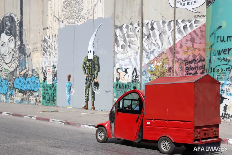 Palestinians walk past a graffiti mural depicting a boy holding a fishbowl looking at a uniformed Israeli soldier wearing a white hood in the style of the Ku Klux Klan (KKK), drawn by Palestinian artist Taqi Spateen alon...