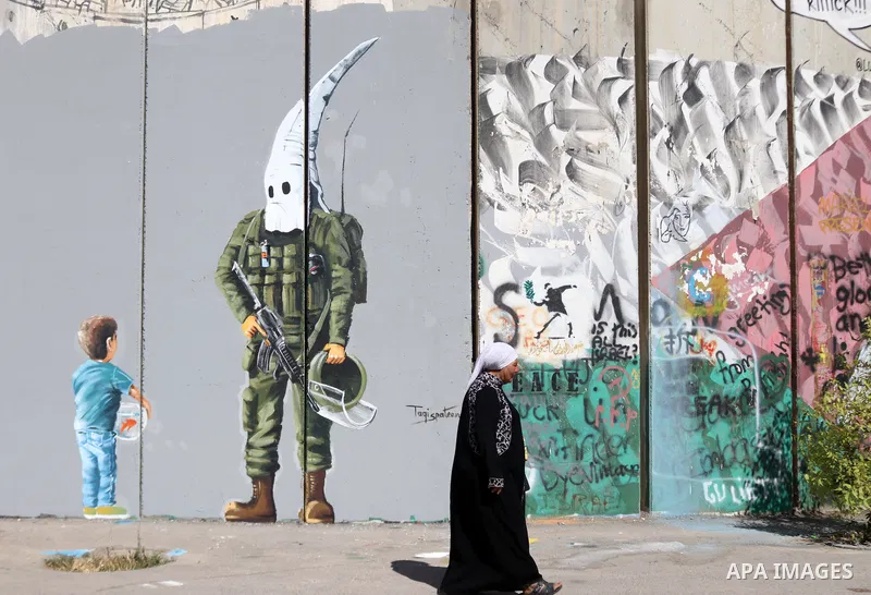 A Palestinian woman walks past a graffiti mural depicting a boy holding a fishbowl looking at a uniformed Israeli soldier wearing a white hood in the style of the Ku Klux Klan (KKK), drawn by Palestinian artist Taqi Spat...